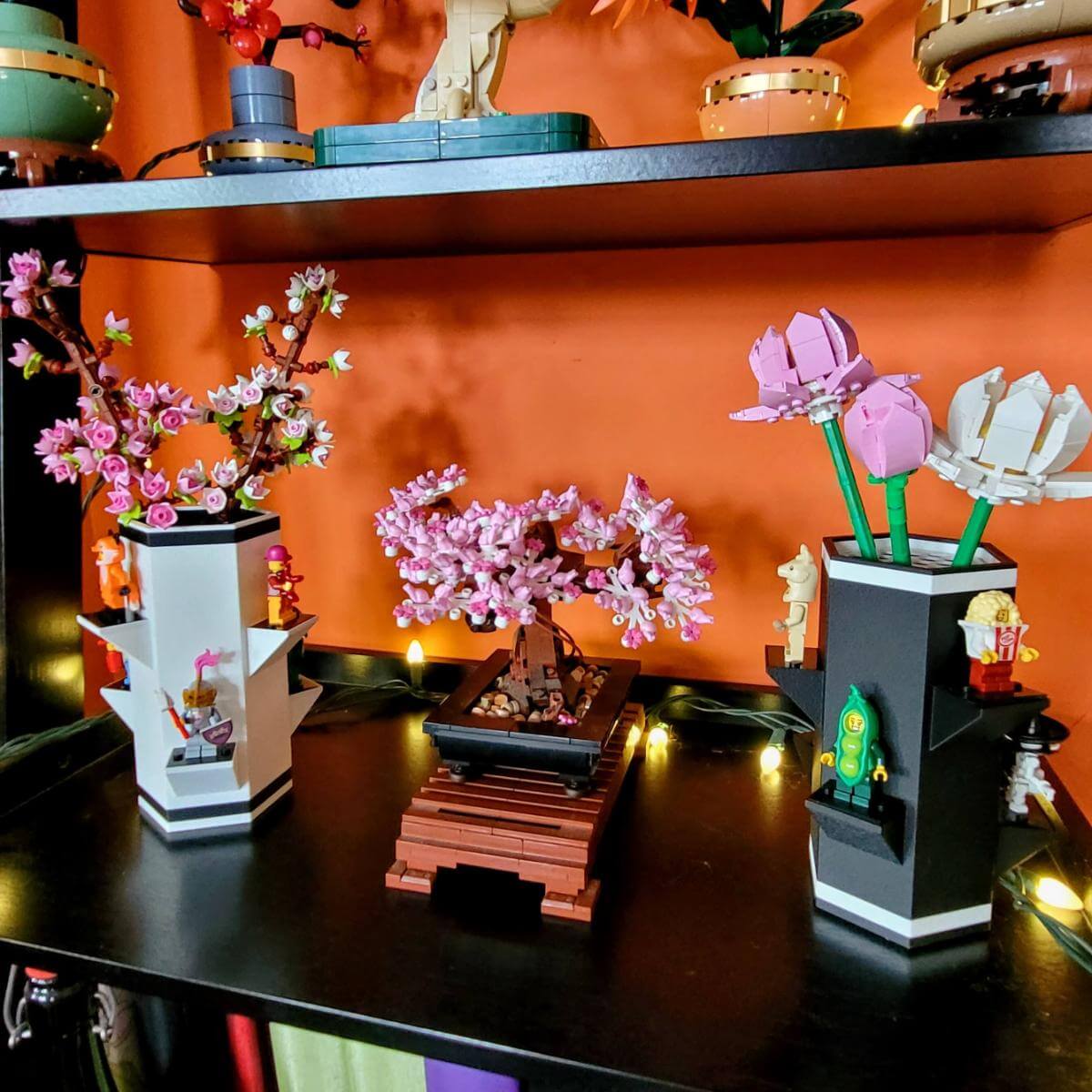 A LEGO botanical flower vase with minifigure display sits on a black shelf, surrounded by other floral LEGO sets in a cozy, warmly lit living space. Features removable platforms for minifigures and compatibility with LEGO flower stems.